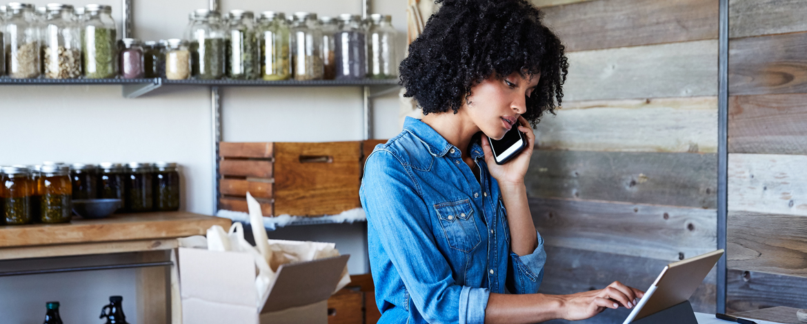 A business woman wearing a denim collared shirt talking on a cell phone and typing on a tablet